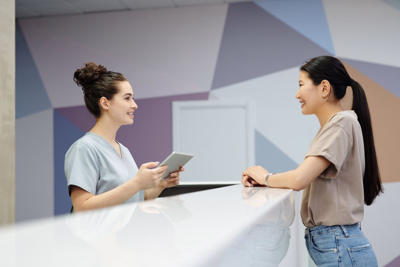 A clinic receptionist holding a tablet while assisting a visitor at a front desk